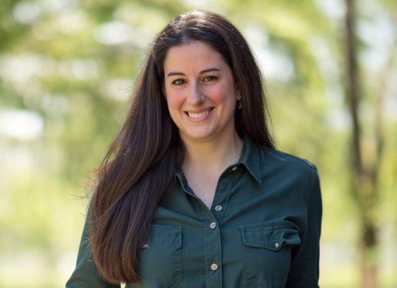 A woman with long dark hair, wearing a dark green button-up shirt, smiles while standing outdoors with blurred greenery in the background.